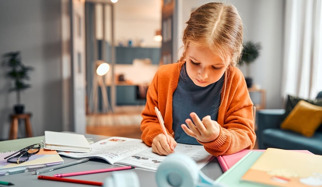 A young girl sits at a table, focused on her notebook while holding a pencil, engaged in writing or drawing.