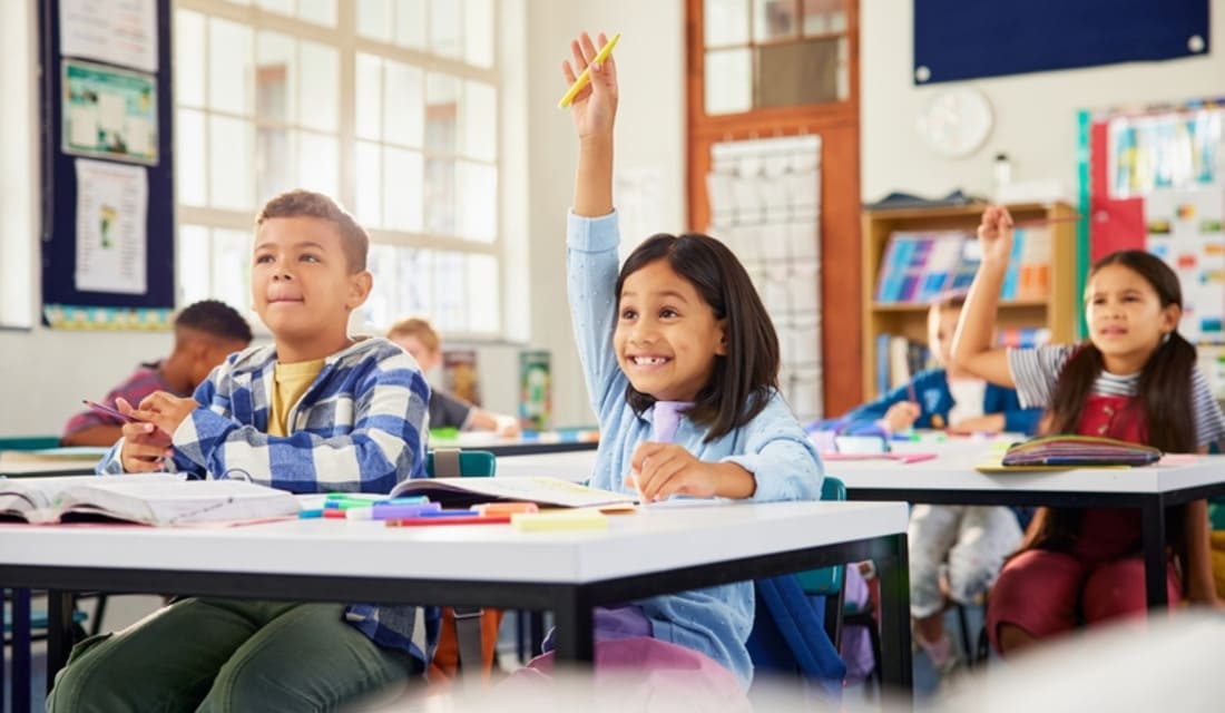Children sitting at desks in a classroom, eagerly raising their hands to answer questions.