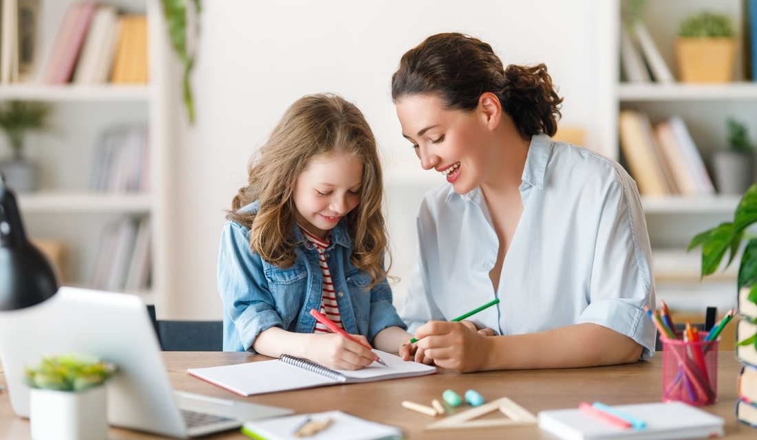 A woman and a child sit together at a desk, focused on a laptop screen in a cozy workspace.